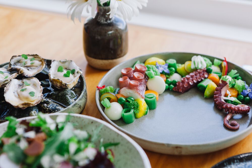 Three plates of food and a small ceramic vase sit on a wooden table. The food includes greens, skinned radishes, and what looks like octopus tentacles.