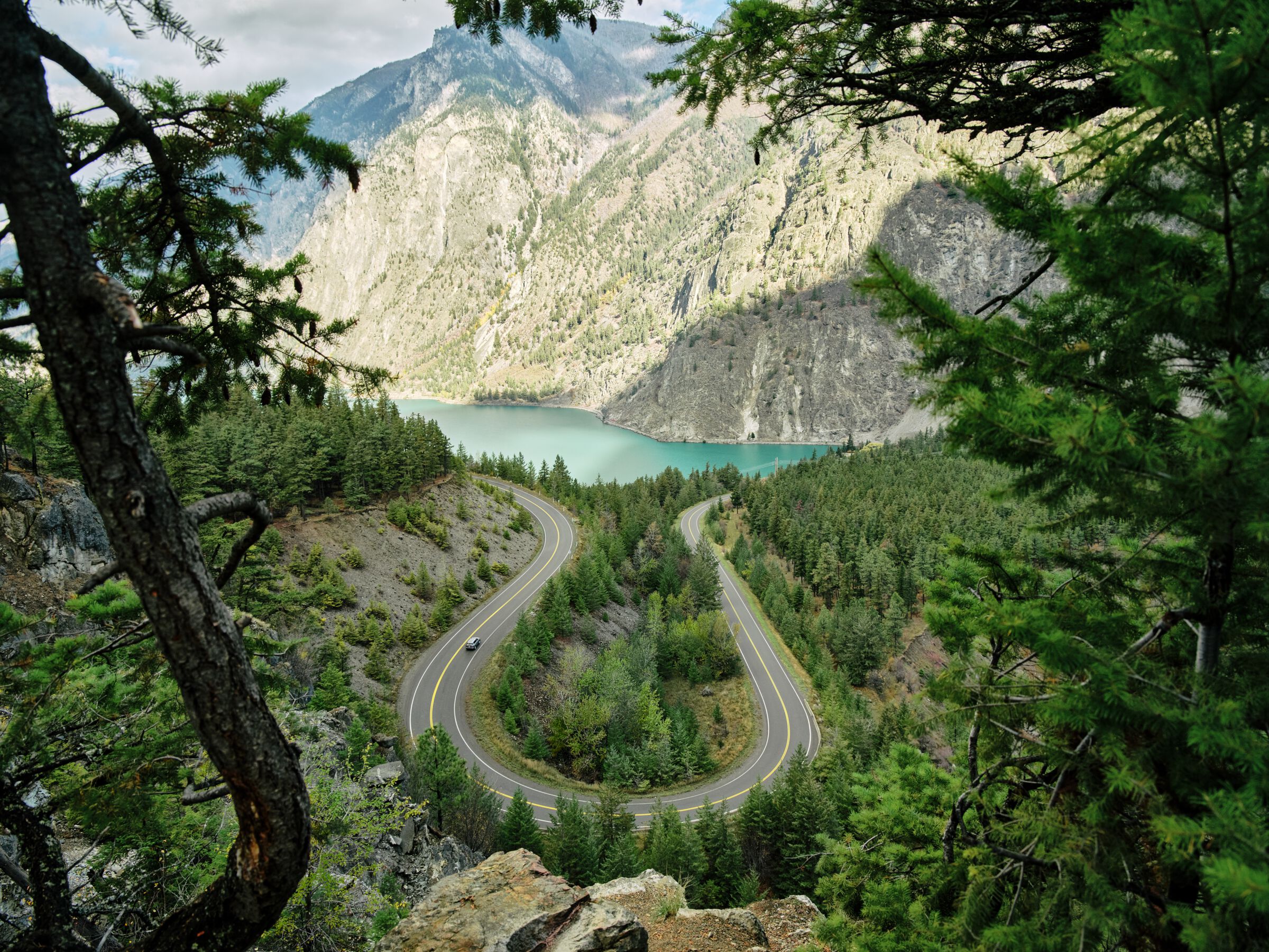 Vehicle travelling along a snaking highway with a turquoise lake and towering mountains in the background.
