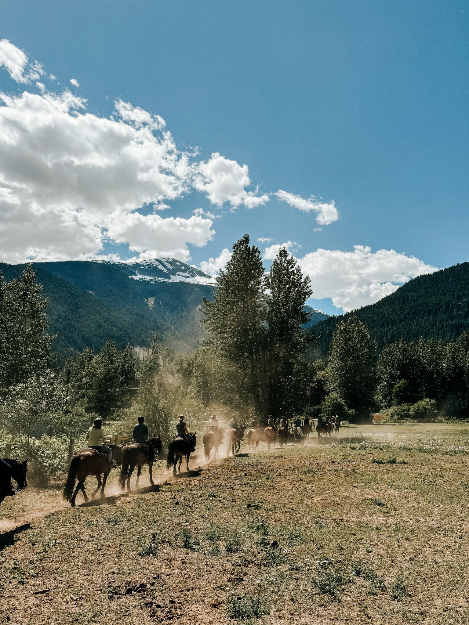 A group of people on horseback walk along a dirt trail with mountains in the background.