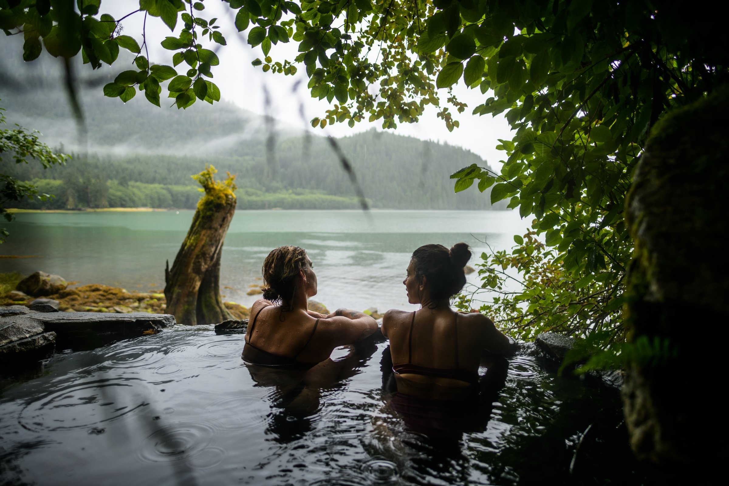 Two people soak in a natural hot springs pool with misty rainforest, mountains and waterways in the distance.