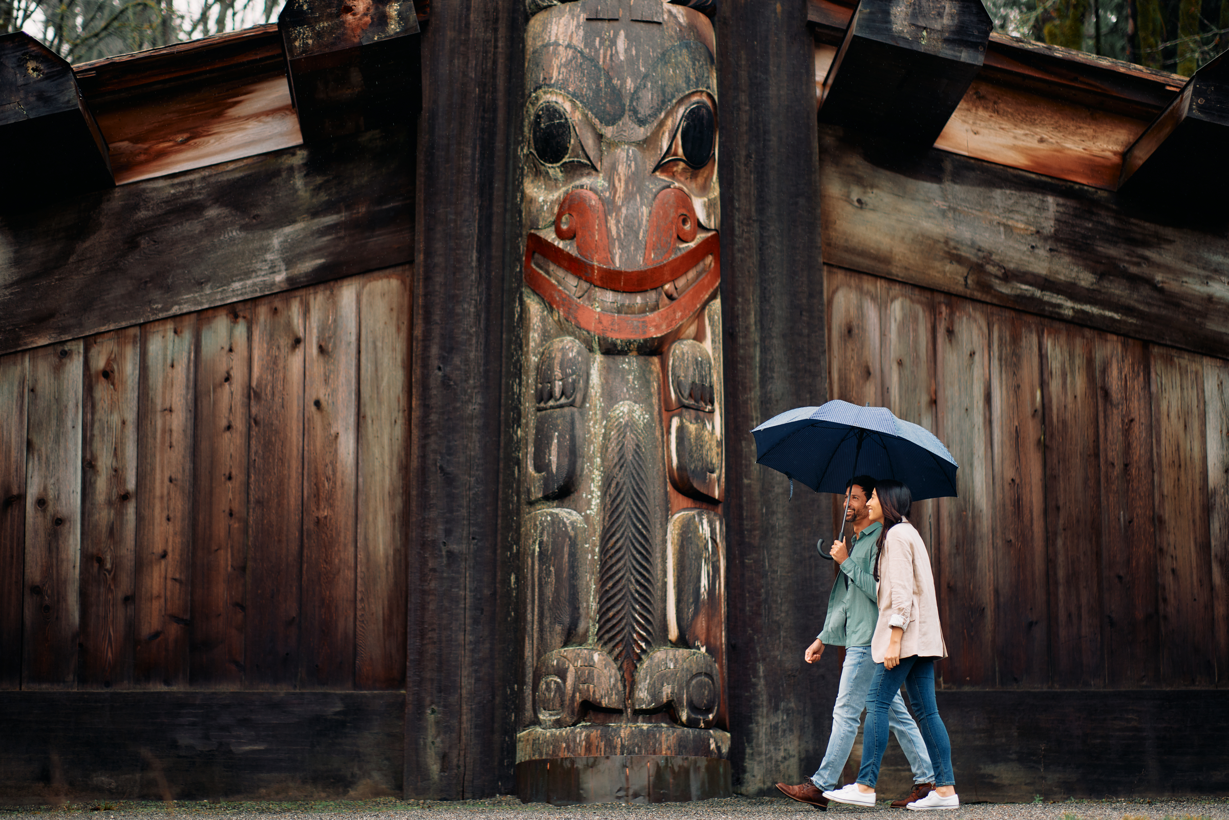 Two people walk under a blue umbrella with a weathered Indigenous pole and building behind them.
