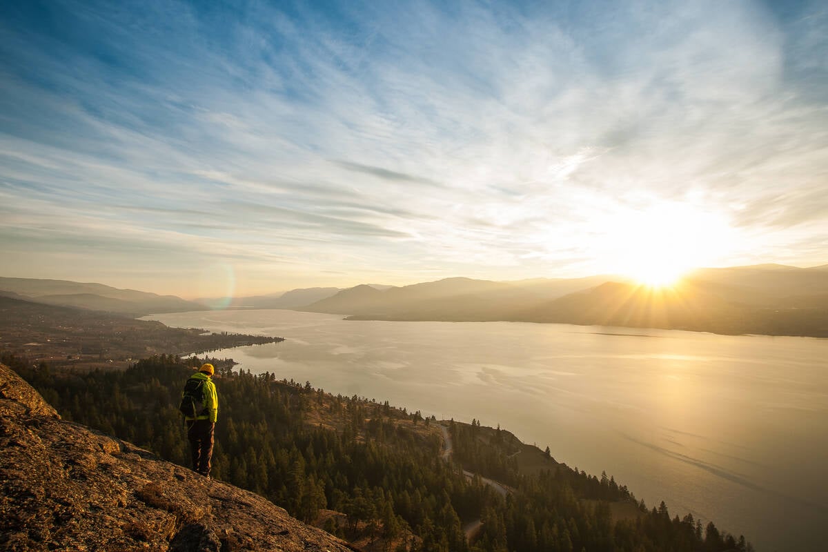 A person stands on a rocky bluff overlooking the lush Okanagan Valley and the large lake as the sun sets on the horizon