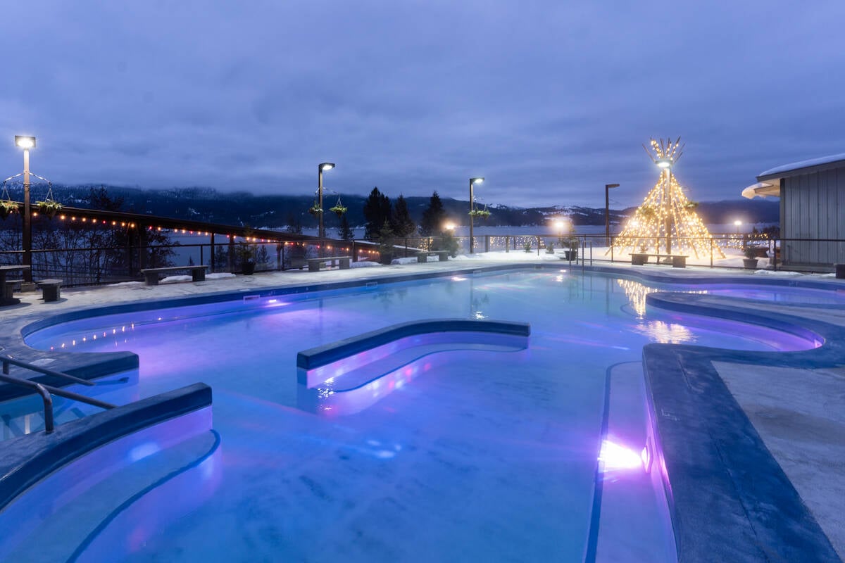 The rounded edges of an empty hot springs pool at dusk. Lights illuminate a teepee structure at the far end of the pool, and in the distance there is a lake and some trees. The sky is cloudy.