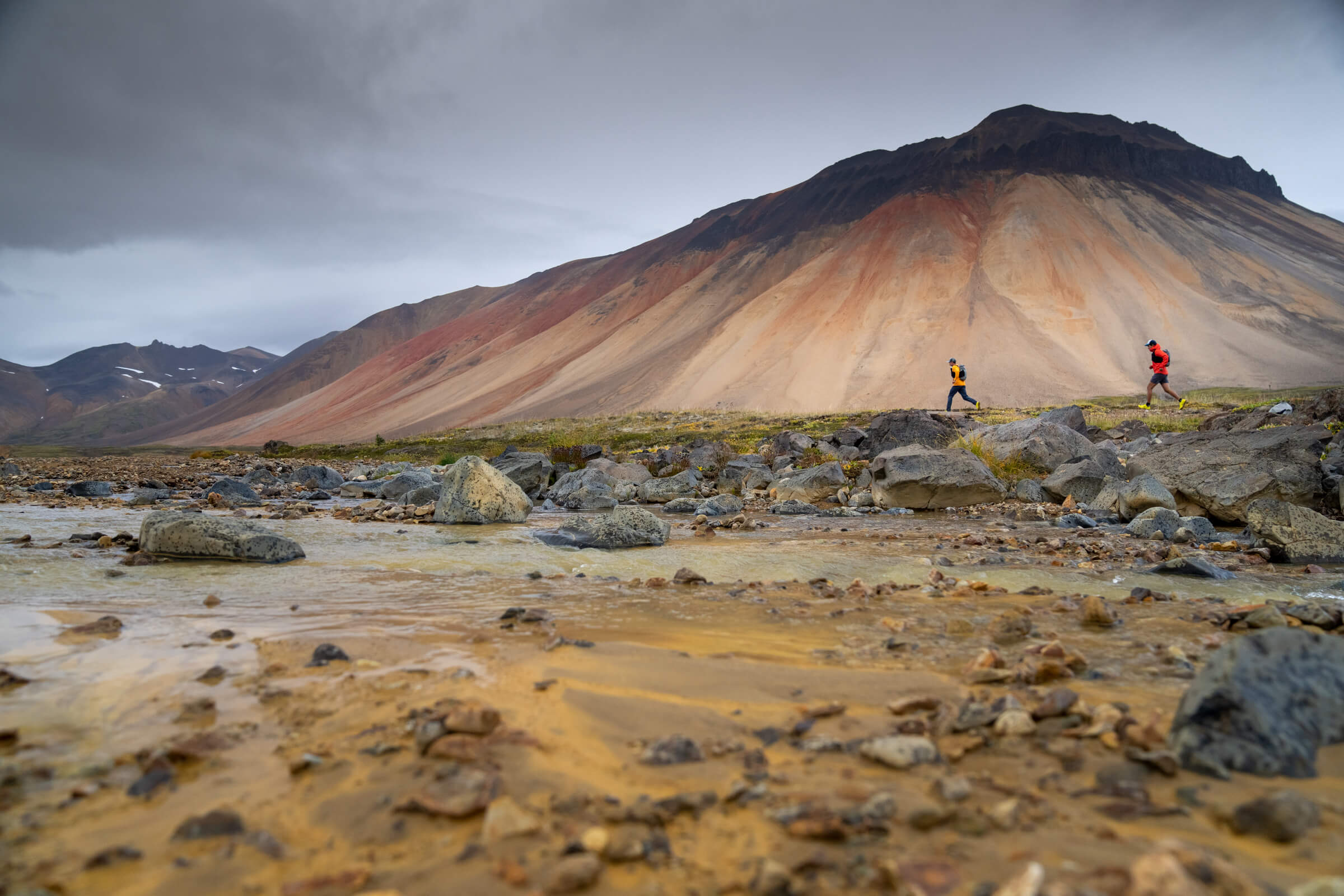 Two trail runners look tiny against the backdrop of a towering Mt. Edziza,streaked with rust and bone colours.