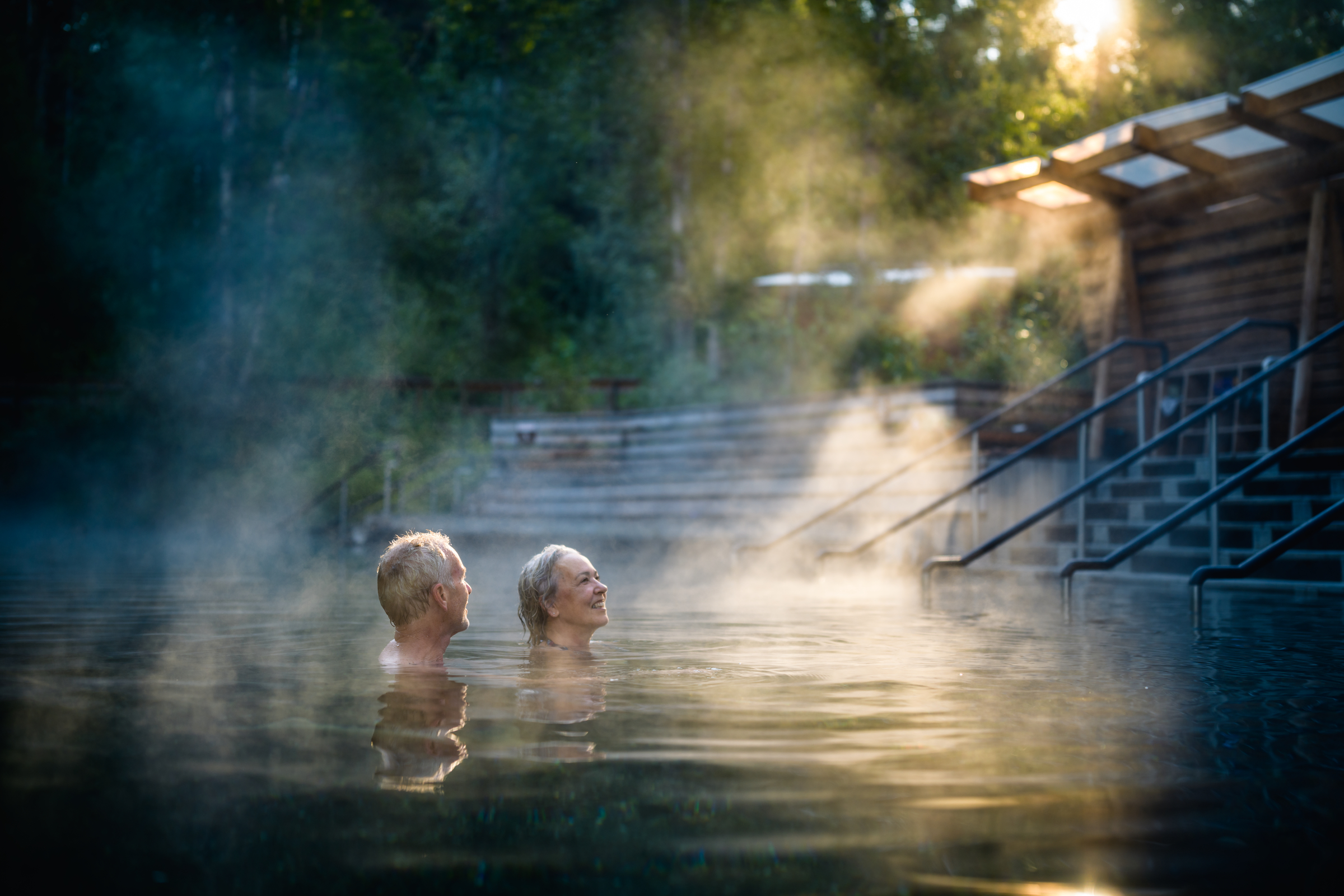 A couple soaks in hot springs with steam rising from the pools as sun peeks through the trees.