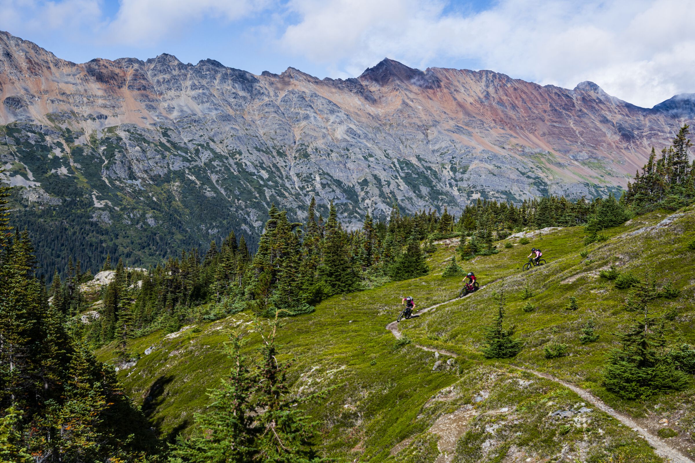 Riders weave their way through a green alpine meadow with jagged peaks in the background.