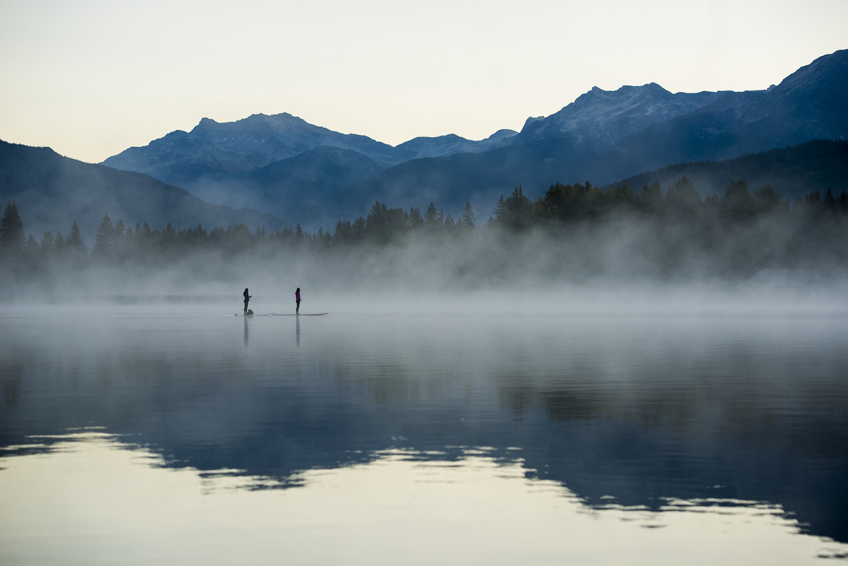 Two paddlers are out on a foggy lake in the morning. Mountains frame the background.