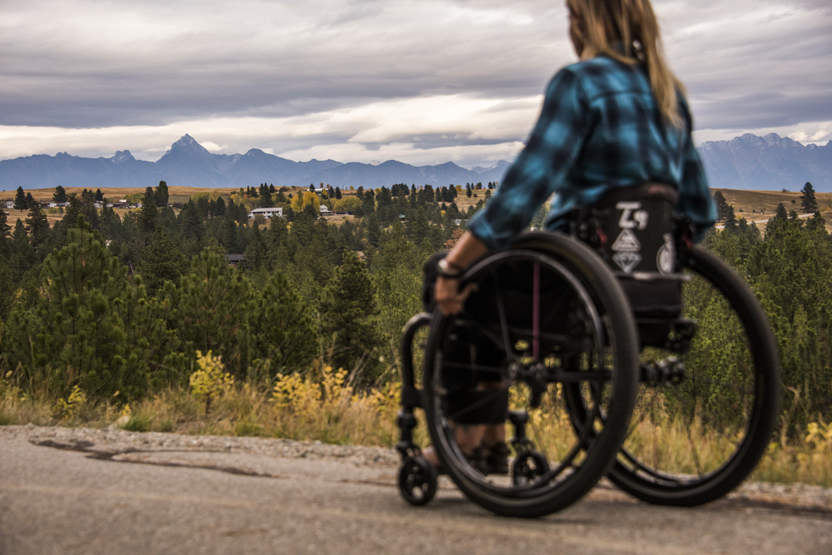 A wheelchair user wearing a blue and black plaid shirt, is on a trail, looking out into the distance. There are evergreen trees and a mountain in the background.