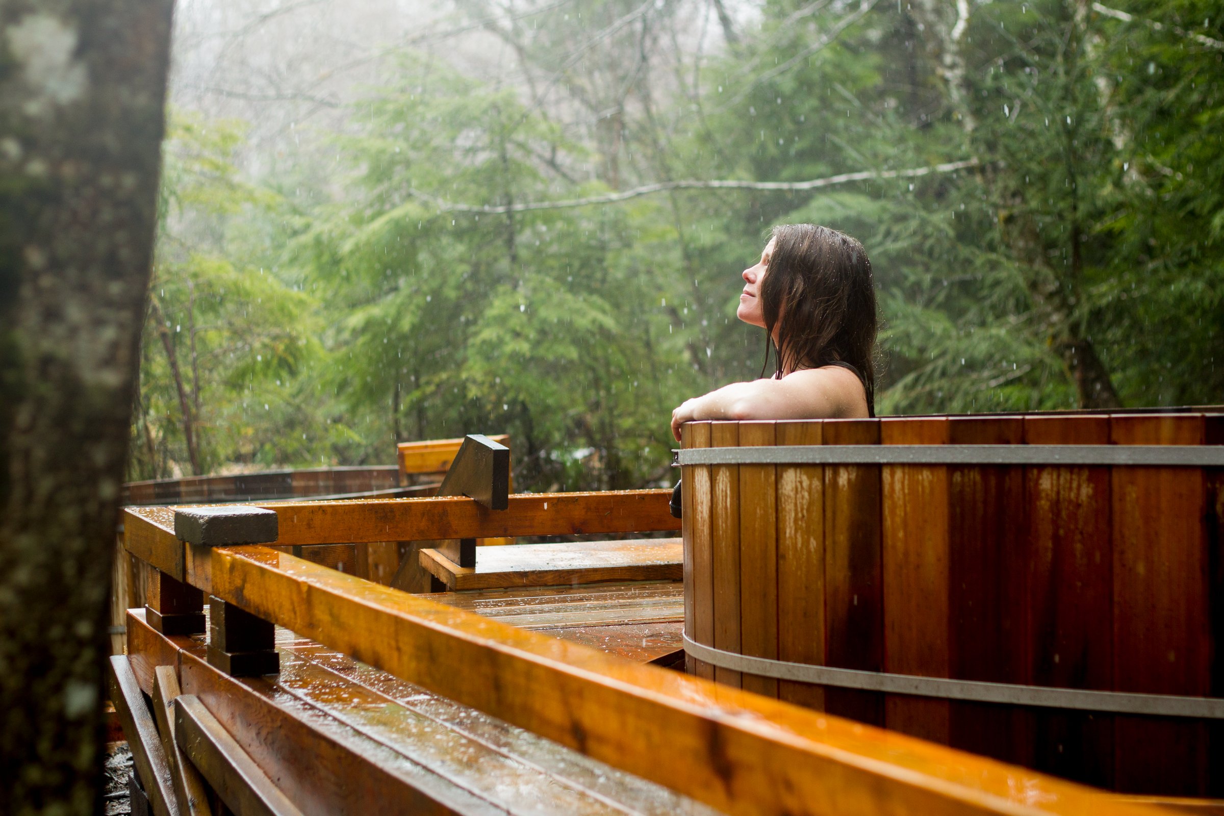 A woman soaks in a wooden tub looking out at the forest.