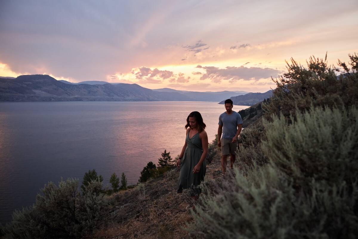 Two people hike along a sagebrush-covered hill above a large body of water with mountains in the distance at sunset