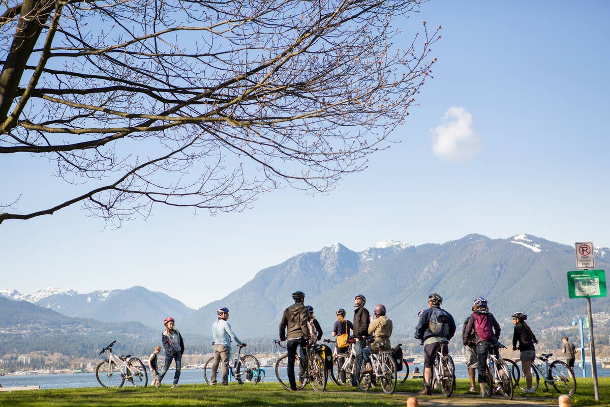A group of a dozen or so cyclists look across a body of water toward the North Shore Mountains on a clear day.