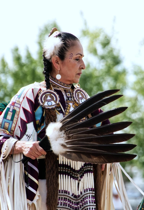 A dancer from Leona Smith - Muskwa River Fancy Dancers - Fort Nelson First Nation, dressed in vibrant traditional attire holds a feather fan.