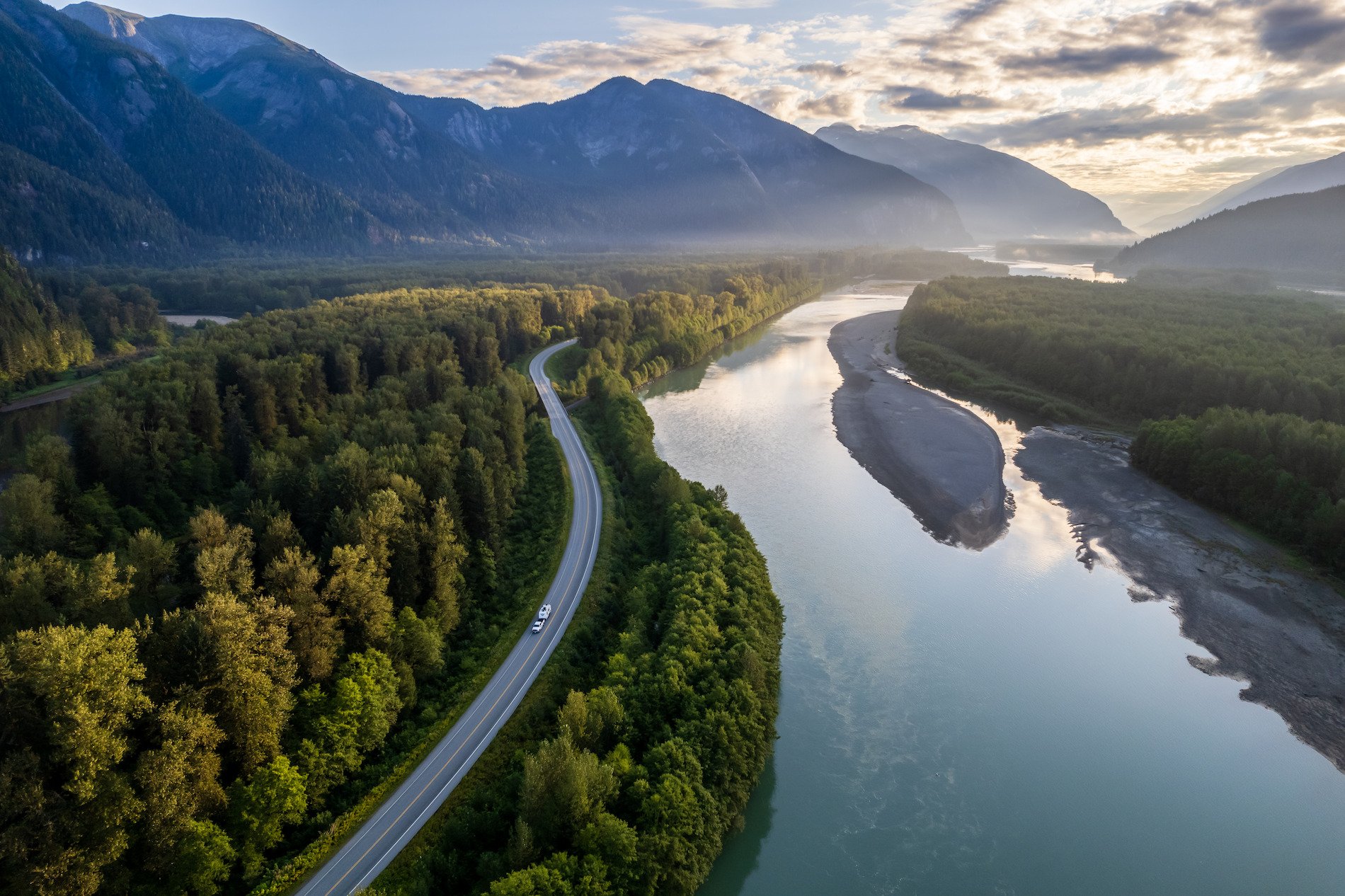 An RV winds its way along a highway, surrounded by dense forest, tall mountains and a calm river.