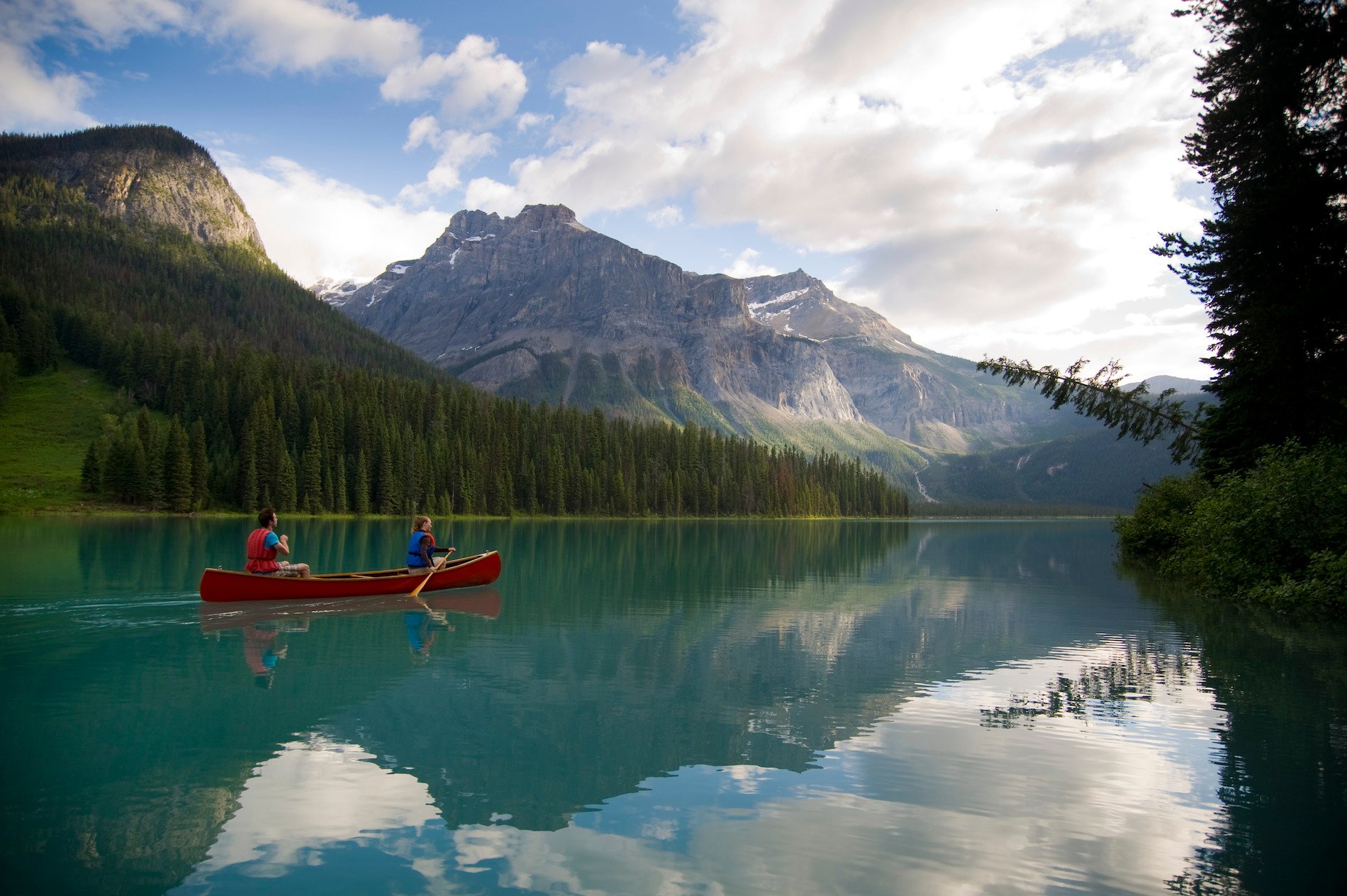 A red canoe is paddled by two people across a lake in Yoho National Park. The turquoise lake is surrounded by mountains and trees.