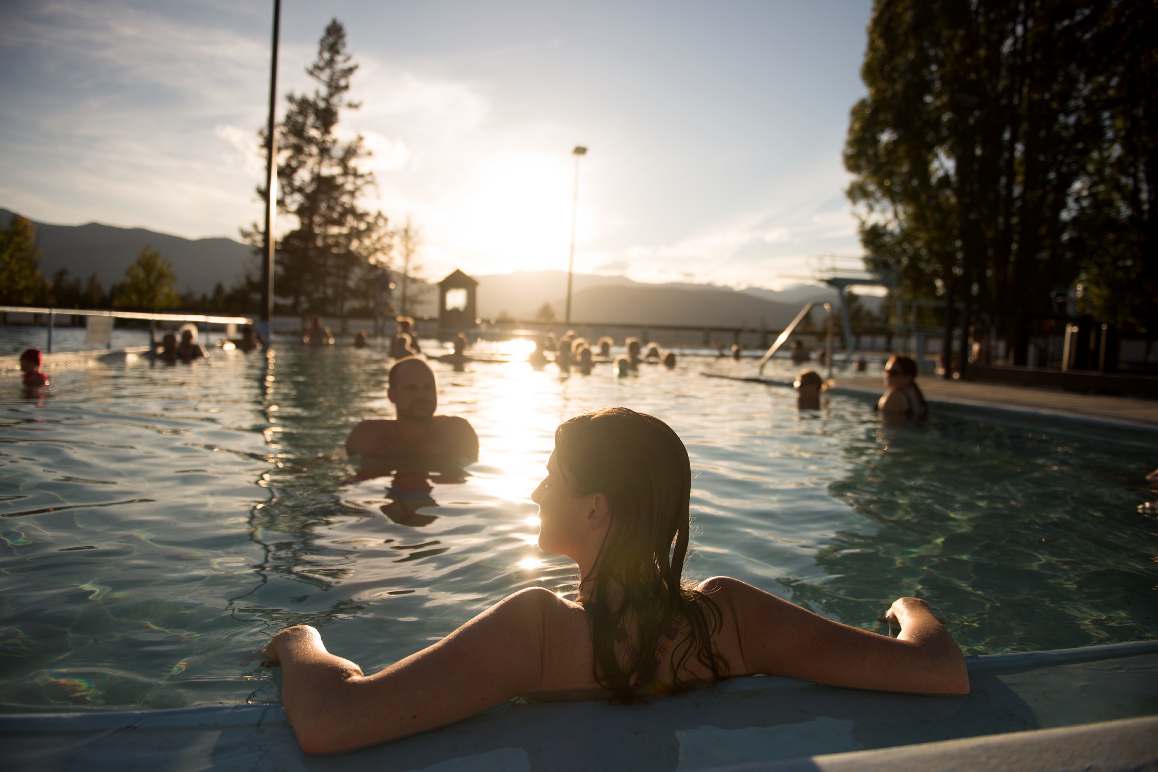 People enjoy a dip in then Fairmont Hot Springs outdoor hot pools. The sun is setting and reflecting across the water. A women in the foreground relaxes by the pool edge with her back to the camera.