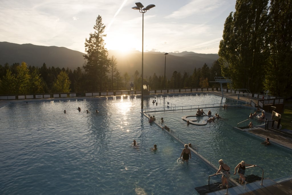 An aerial shot of a large hot springs pool with 20 or so people in the water. In the distance the sun is setting over the mountains and there are some evergreen trees just beyond the pool.