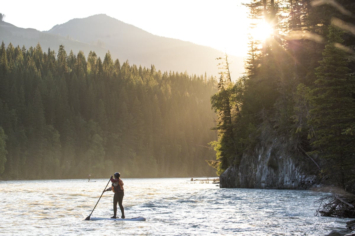 A paddleboarder navigates a serene river surrounded by dense forests, with the setting sun casts a warm golden light.
