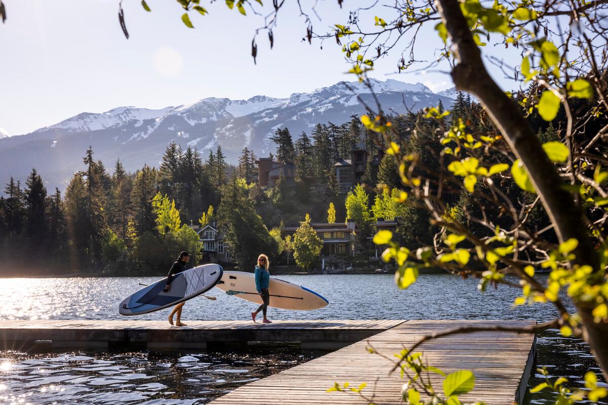 Two people carrying paddleboards on the dock of a lake with houses on the shore behind them.