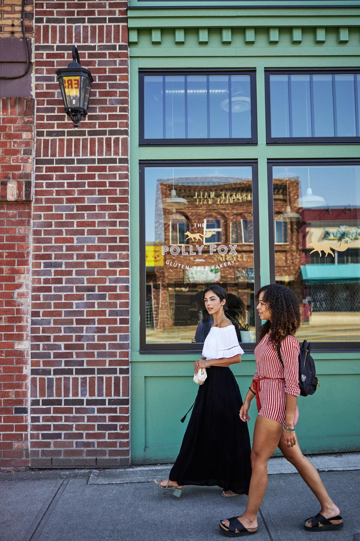 Two friends walking on a sidewalk in front of a bakery storefront.