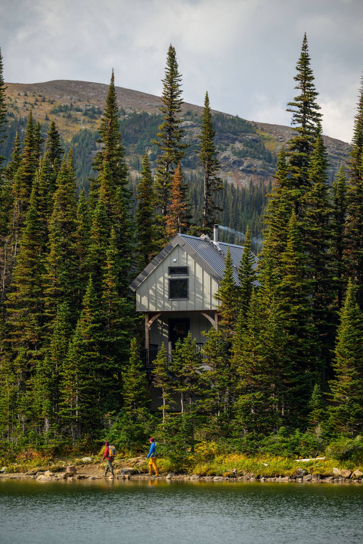 A quaint cabin is tucked in a forest overlooking a lake with towering mountains in the background.
