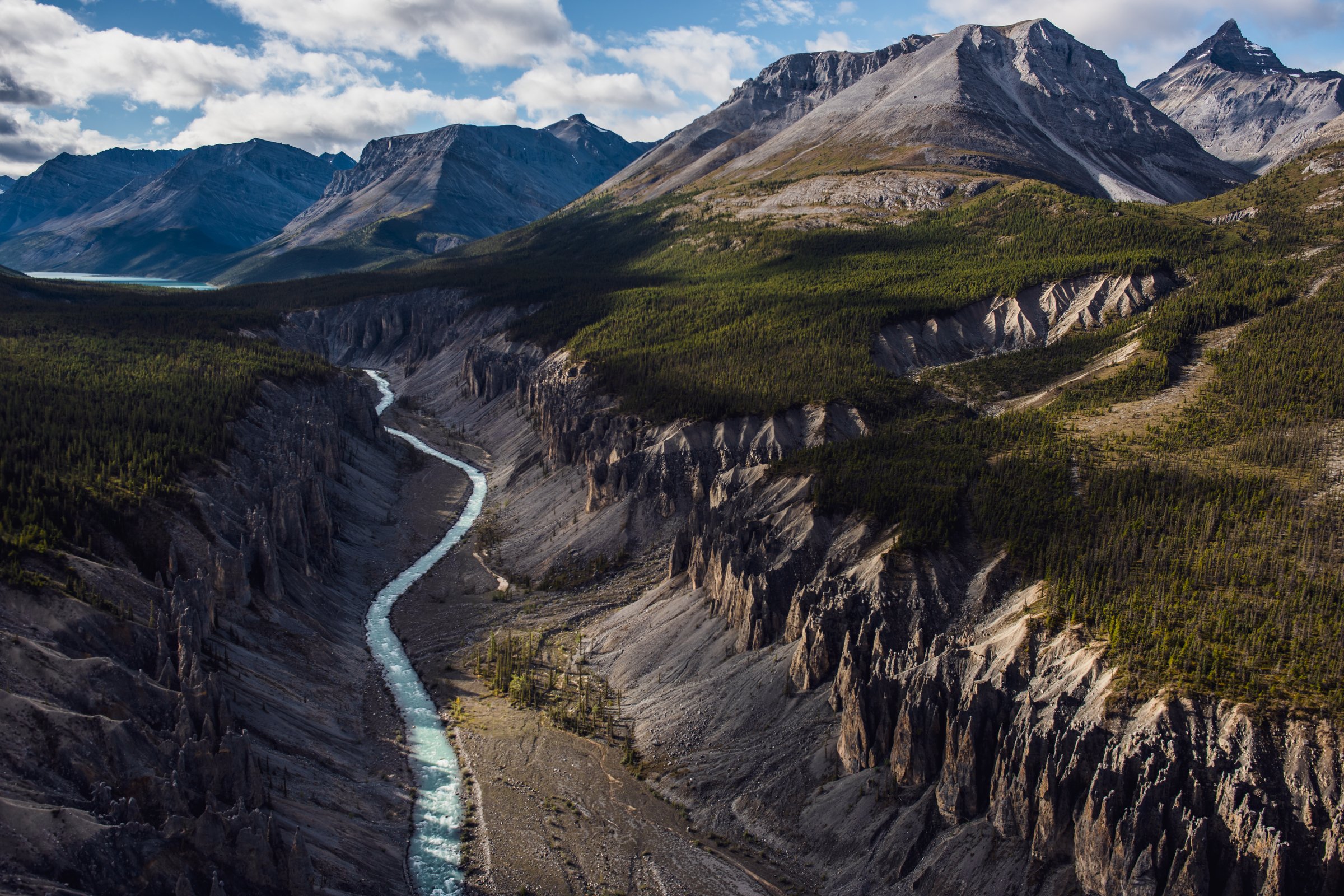 A torquoise river slices through a canyon edged by rocky cliffs, forested foothills and towering mountains in the background.