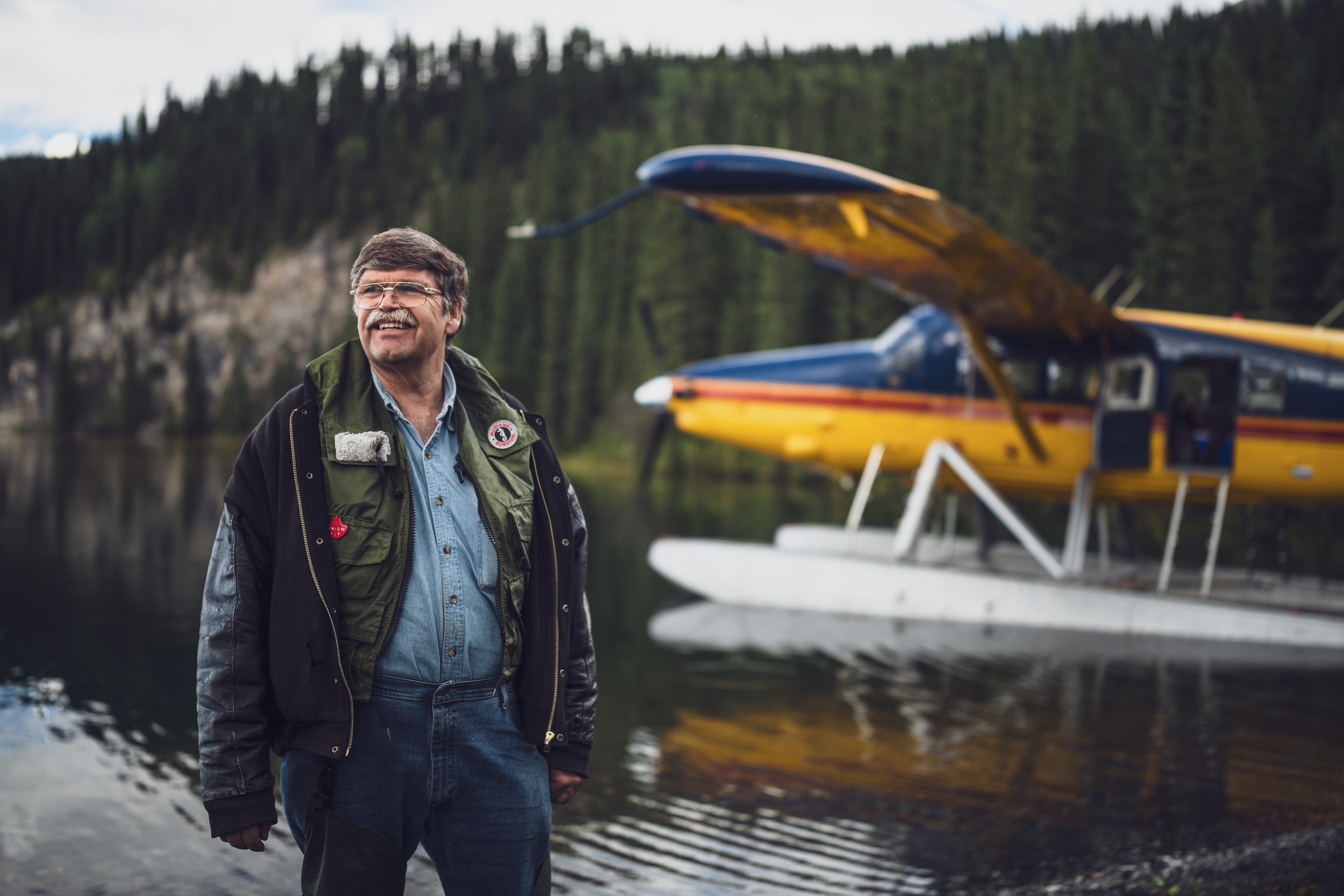 A rugged looking pilot walks beside his float plane, which has landed on a remote lake.